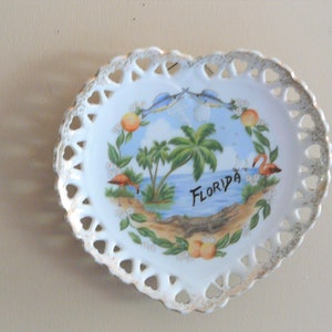Könnte beinhalten: Weiße herzförmige Keramikplatte mit einer spitzengleichen Umrandung. Die Platte zeigt eine bemalte Szene mit Palmen, Flamingos, Fischen und dem Wort "Florida".