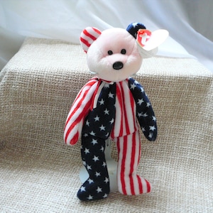 May include: A pink and white beanie baby bear wearing a red and white striped shirt with blue stars and a blue and white striped shirt with red stars. The bear has a red and white tag attached to its ear.