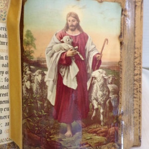 May include: A vintage religious book with a gold cover and a velvet ribbon bookmark. The cover features a picture of Jesus carrying a lamb with a flock of sheep in the background.