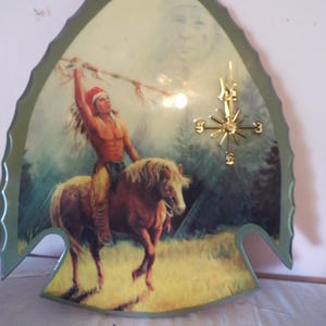 May include: An arrowhead-shaped wall clock featuring a printed illustration of a person in a feathered headdress riding a horse while holding a spear. The clock face includes a gold-colored compass-style dial with cardinal direction markers.