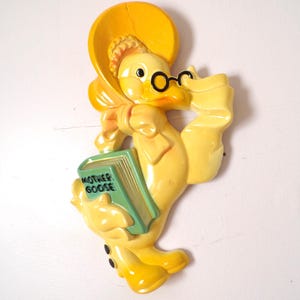 May include: A yellow duck figurine wearing a hat and glasses, holding a green book that reads "Mother Goose." The duck has a bow and black accents. The figurine is a vintage decorative item.