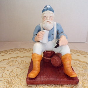 May include: A porcelain figurine of a man wearing a blue and white hat, a blue shirt, white pants, and orange boots. He is sitting on a wooden barrel and holding a white mug.