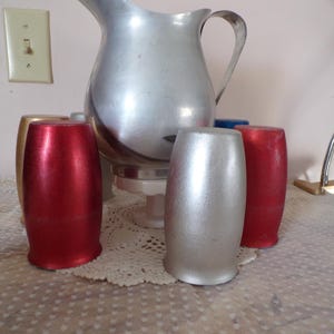 May include: A vintage aluminum pitcher with a handle and spout, accompanied by several metallic tumblers in red, silver, and gold. The pitcher and tumblers are arranged on a lace doily, suggesting a retro aesthetic.