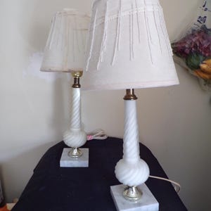 Puede incluir: Par de lámparas de mesa de alabastro blanco con pantallas de lámpara con flecos blancos. Las lámparas tienen un diseño retorcido y se apoyan sobre bases cuadradas de mármol.