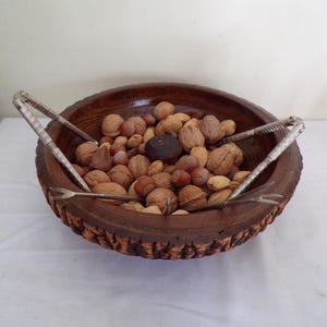 May include: A wooden bowl filled with various nuts, including walnuts, hazelnuts, and almonds. Two silver nutcrackers and a pick are placed on the bowl's rim. The bowl has a textured, dark brown exterior.