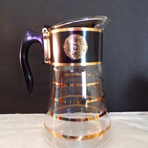 Puede incluir: Una jarra de vidrio vintage con una banda negra con un diseño de medallón dorado. La jarra tiene un asa negra y está adornada con rayas doradas. El vidrio transparente permite la visibilidad del contenido. Una pieza clásica para servir bebidas.