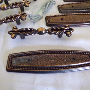 May include: Bronze-toned cabinet hardware set, including ornate handles and rectangular backplates. The handles feature a twisted rope design with decorative ends. Screws are included for installation. The hardware is designed for furniture.
