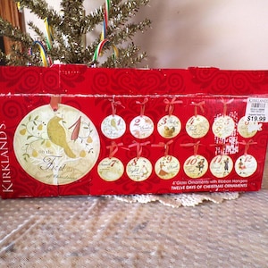 May include: A red box of Kirkland's Christmas ornaments. The box features a large circular ornament with a bird and the text "on the First day of Christmas." The box contains 12 glass ornaments with ribbon hangers.
