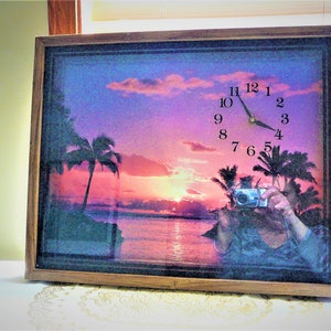 Puede incluir: Un reloj enmarcado con una escena de puesta de sol con palmeras y una playa. La esfera del reloj se superpone a la escena y muestra la hora 1:45. El reloj está en un marco de madera.