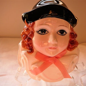 May include: A ceramic figurine of a woman with red hair wearing a black hat and a pink bow. The figurine is wearing a white ruffled collar and has a painted face with blue eyes.