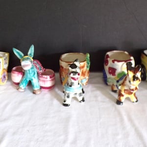 Puede incluir: Una colección de figuras de animales de cerámica, incluyendo burros y caballos, con detalles coloridos. Algunas figuras están emparejadas con macetas o carretas, mostrando una estética caprichosa y vintage. Las piezas están pintadas a mano con colores brillantes.