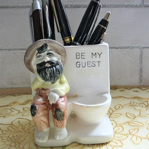 Pode incluir: Porta-canetas de cerâmica branca em forma de um homem sentado em um vaso sanitário. O homem tem barba e usa um chapéu. O vaso sanitário tem a frase "Be My Guest" escrita nele.