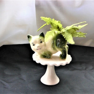 Può includere: Un vaso da fiori in ceramica bianco e verde a forma di gatto con una pianta di felce verde al suo interno. Il vaso è posto su un piedistallo per torte bianco.
