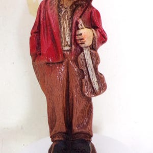May include: A carved wooden figurine of a smiling man in a red coat, brown pants, and a blue top hat. The figure is holding a violin. The figurine is standing on a small wooden base.