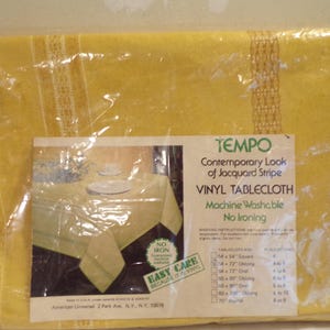May include: A yellow vinyl tablecloth with a jacquard stripe design, packaged in clear plastic. The label reads "TEMPO Vinyl Tablecloth" and includes a picture of the tablecloth on a table. It is machine washable and requires no ironing.