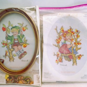 May include: Two oval-shaped cross-stitch embroidery kits featuring children in trees. The kits are packaged in plastic bags with instructions and floss. The first kit features a boy in a green shirt and brown shorts, sitting on a branch with red apples. The second kit features a girl in a red dress and blue apron, sitting on a branch with red berries.