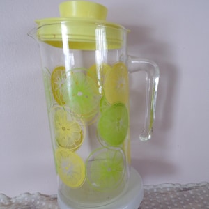 Puede incluir: Una jarra de vidrio transparente con tapa y asa amarillas, decorada con rodajas de cítricos amarillos y verdes. La jarra está sobre un pedestal blanco. Una adición refrescante a cualquier cocina.