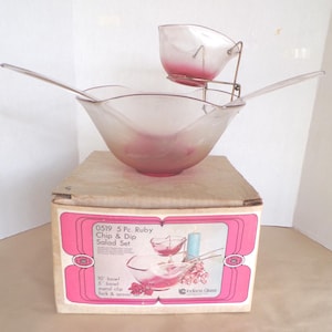 Op de afbeelding: Een vintage 5-delige ruby chip- en dipsaladeset. De set bevat een grote kom, een kleinere kom op een metalen standaard en een vork en lepel. De kommen hebben een roze tint. De set zit in de originele doos.