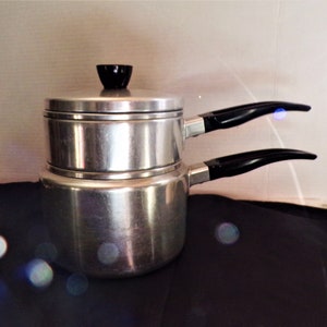 Puede incluir: Un juego de dos ollas de aluminio plateado con asas y tapas negras. Las ollas están apiladas una encima de la otra.