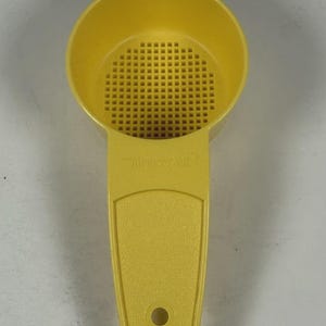 Puede incluir: Un colador de plástico amarillo con un mango. El colador tiene un cuenco redondo con una rejilla de pequeños agujeros cuadrados. El mango tiene una superficie texturizada y un agujero en el extremo. La marca es visible en el mango.