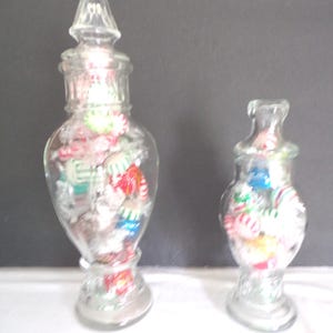 May include: Two clear glass candy jars filled with colorful candies. The taller jar has a pointed lid, while the shorter jar has a rounded lid. Both jars are filled with red, green, white, and blue candies.