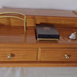 Puede incluir: Un organizador de escritorio de madera con dos cajones con pequeños pomos dorados, un arco de metal dorado, una caja negra rectangular con un emblema de estrella en la tapa y un anillo plateado en una pequeña bandeja empotrada a la derecha.