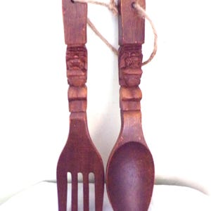 以下が含まれることがあります： スプーンとフォークの形をした木製壁飾りセット。ハンドルはティキの顔で彫刻されており、食器は濃い茶色に塗られています。