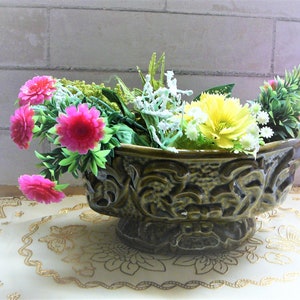 May include: A green ceramic planter with an ornate design, filled with pink and yellow artificial flowers. The planter sits on a white lace doily.
