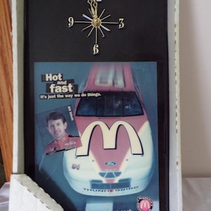 May include: A rectangular wall clock featuring a black face with gold-toned hands and numerals at 3, 6, 9, and 12. The lower section displays a Ford Thunderbird race car with a McDonald's logo, a portrait of Bill Elliott, and the text Hot and fast.