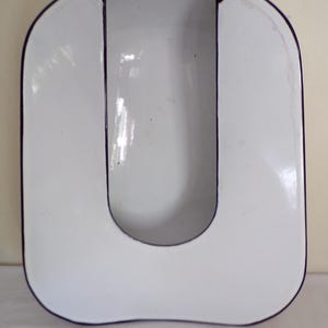 May include: A white, U-shaped enamel bedpan with a dark blue trim. The bedpan has a smooth, glossy finish and a rectangular shape with rounded corners. The interior is also white, and the opening is at the top.