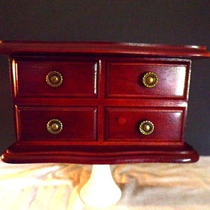 Puede incluir: Pequeña caja de joyería de madera rojo oscuro con cuatro cajones, cada uno con un pomo dorado. La caja se asienta sobre una base blanca tipo pedestal. La madera tiene un acabado brillante.