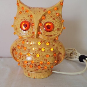 May include: A ceramic owl-shaped lamp with a mottled yellow and brown finish. The owl features two large, glowing orange eyes and numerous small, illuminated orange plastic studs embedded across its body, connected to a white electrical cord.