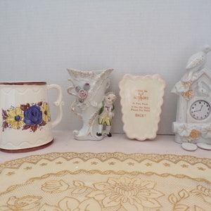 May include: A set of four vintage porcelain figurines. The figurines include a watering can with floral designs, a vase with a floral design, a small figurine of a boy, and a clock with a bird on top. The figurines are all white with gold accents.