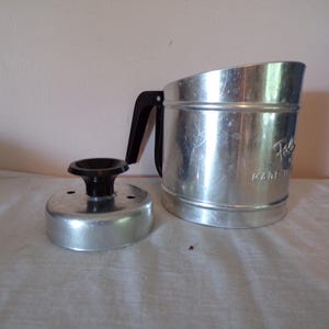 May include: A silver-colored metal flour sifter with a black plastic handle and a separate matching metal lid featuring a black plastic knob. The side of the sifter body has embossed text that reads Foley and Made in USA.