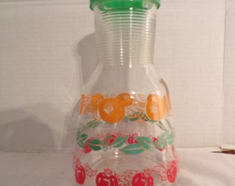 May include: A clear glass pitcher with a green lid and a fruit design. The design features orange and red fruits with green leaves.