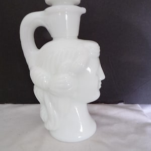 Könnte beinhalten: Weiße Keramikflasche mit einem Frauenkopfdesign. Die Flasche hat einen Griff und einen dekorativen Deckel.