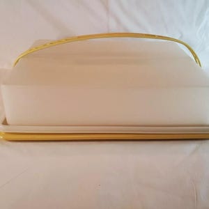 May include: A rectangular cake carrier featuring a translucent white plastic lid and a solid yellow plastic base. The container includes a matching yellow arched handle that spans across the top of the lid for secure transport.