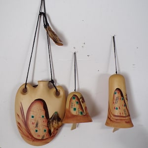 May include: Hand-painted, beige ceramic wind chimes with a Southwestern theme. The set includes a flask-shaped piece and two bell-shaped chimes, all suspended by black cords. Each piece features a painted landscape with dwellings and geometric patterns.