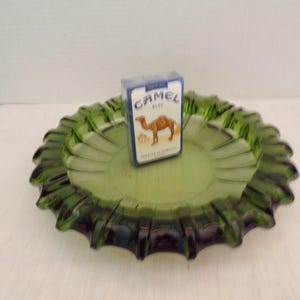 Puede incluir: Un cenicero de vidrio verde con un borde festoneado. Un paquete de cigarrillos Camel está sentado encima del cenicero.