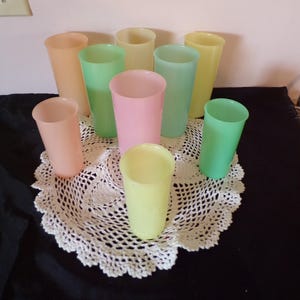 May include: A collection of pastel-colored plastic tumblers in shades of peach, green, pink, and yellow. The cups are arranged on a white, crocheted doily, set against a dark background. The tumblers are cylindrical and appear to be vintage.