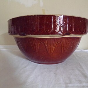 May include: A large, brown ceramic mixing bowl with a white rim and a decorative pattern. The bowl has a scalloped edge and a textured surface.