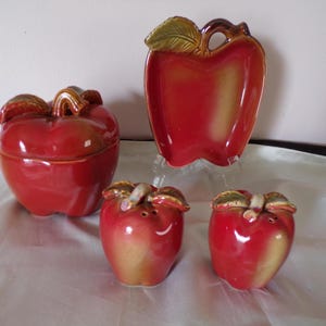 Puede incluir: Un juego de cocina de cerámica con temática de manzana. El juego incluye un tarro con tapa, una fuente para servir y dos saleros y pimenteros. Los artículos son principalmente rojos con detalles marrones y dorados.