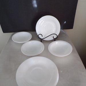 May include: Five white ceramic saucers with a slightly raised center. The saucers are arranged on a white surface.