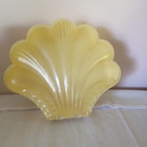 Op de afbeelding: Een lichtgeel, schelpvormig decoratief object. Het object heeft een geschulpte rand en een geribbelde textuur, die op een zeeschelp lijkt. Het item is waarschijnlijk gemaakt van plastic of een vergelijkbaar materiaal. Het object ligt op een wit oppervlak.