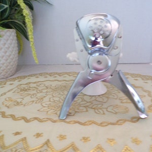 May include: A polished, silver citrus juicer with a perforated top and a hinged mechanism. The juicer has three legs and is placed on a cream-coloured surface with gold floral embroidery. A white ceramic vase and a potted plant are in the background.