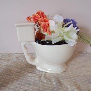 Puede incluir: Un jarrón de cerámica con forma de inodoro, pintado de blanco con un borde negro, lleno de flores artificiales de colores. Las flores incluyen flores naranjas, blancas y azules, creando una pieza decorativa caprichosa.