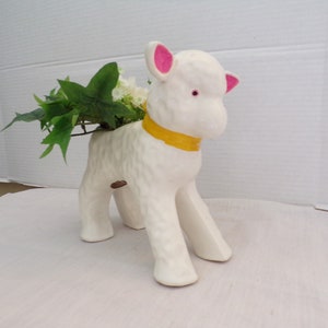 May include: White ceramic lamb planter with pink ears and a yellow ribbon around its neck. The planter is holding a small green plant.