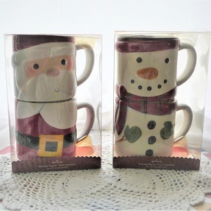 May include: Two sets of Hallmark stackable mugs in clear plastic packaging. One set features a Santa Claus design with a red suit and white beard. The other set has a snowman design with a burgundy scarf and a carrot nose. Each set contains two mugs.