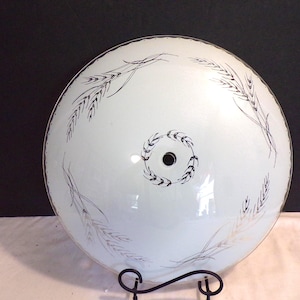 Può includere: Un paralume rotondo in vetro bianco con un motivo a spighe di grano nero e argento. L'apparecchio ha un cerchio nero centrale ed è sostenuto da un supporto in metallo nero. Il motivo del grano è sul bordo dell'apparecchio.