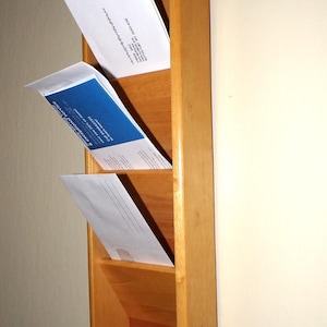 Può includere: Un organizer da parete in legno con tre scomparti angolati per lettere e buste. L'organizer è realizzato in legno chiaro e ha un portachiavi in basso. Diverse buste sono collocate negli scomparti.